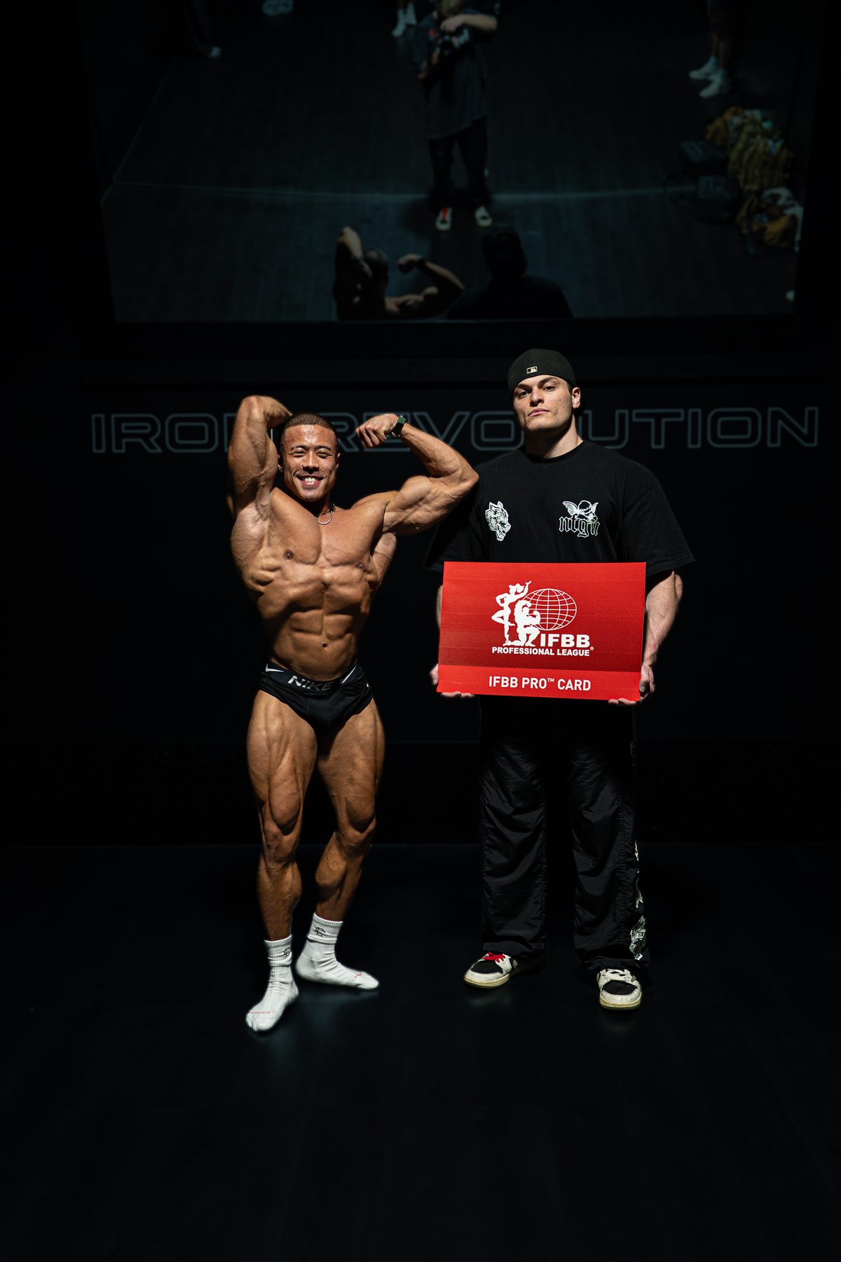 Client flexing beside Jake holding an IFBB Pro Card